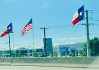 Texas state flags and American flag flying on poles outside a building, symbolizing regional identity and patriotism, photographed from a moving vehicle on a sunny day.