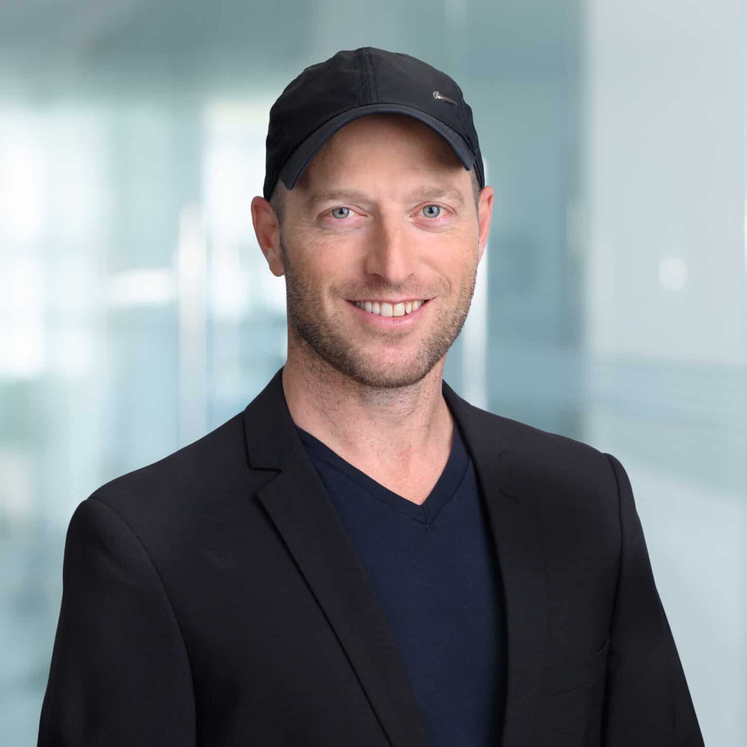 - Young man wearing a black blazer, dark T-shirt, and black cap, smiling confidently, in a modern, bright office environment.